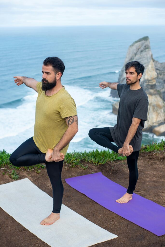 Two men practicing yoga on cliffside in Portugal with ocean view, embracing serenity and fitness.